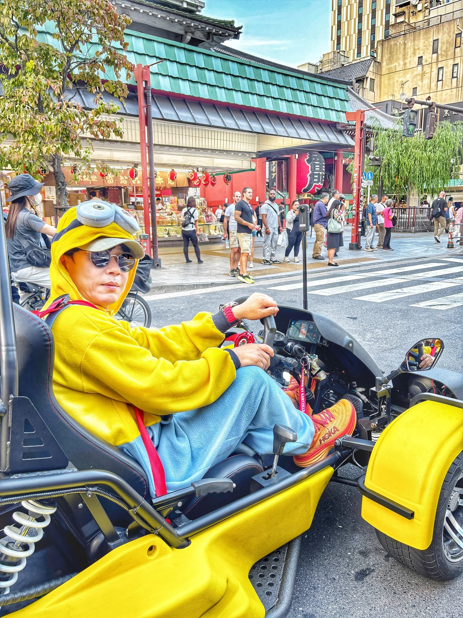 Person in a bright yellow Minion costume steering a vibrant yellow go-kart through Asakusa, with a traditional temple gate in the background.