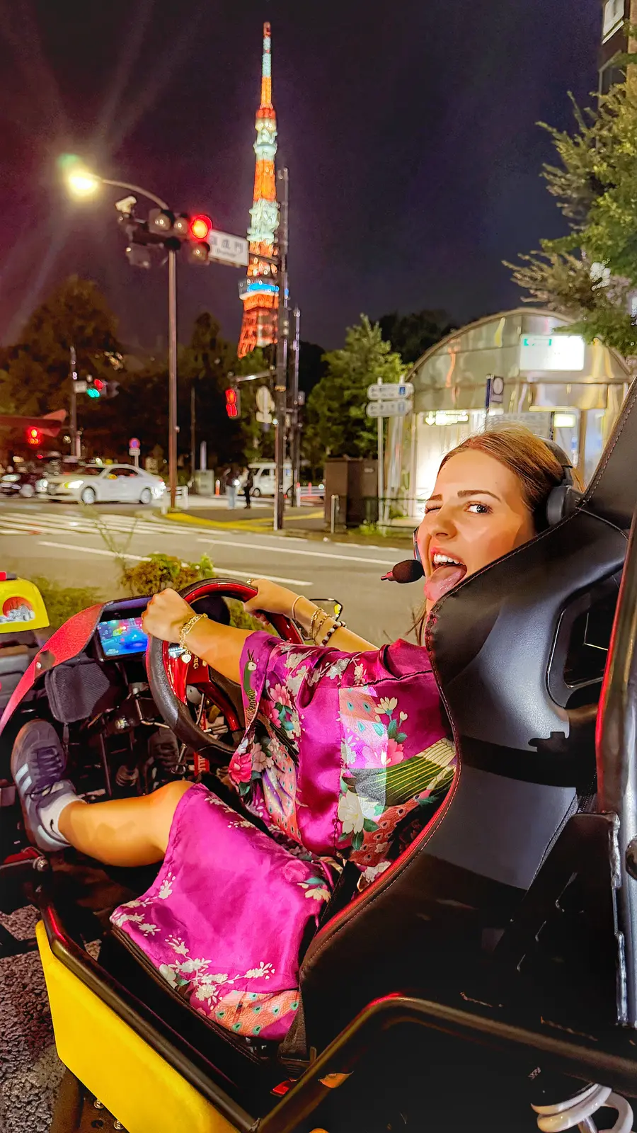 Woman in a pink floral kimono-style robe sticking out her tongue playfully from her yellow go-kart at night, with the glowing Tokyo Tower in the background.