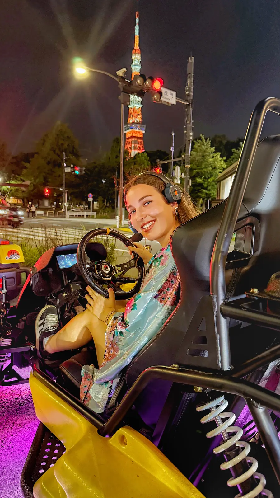 Smiling woman in a mint floral kimono-style robe holding the wheel of her yellow KartZilla go-kart at night, pink underglow glowing, with the illuminated Tokyo Tower rising behind her.
