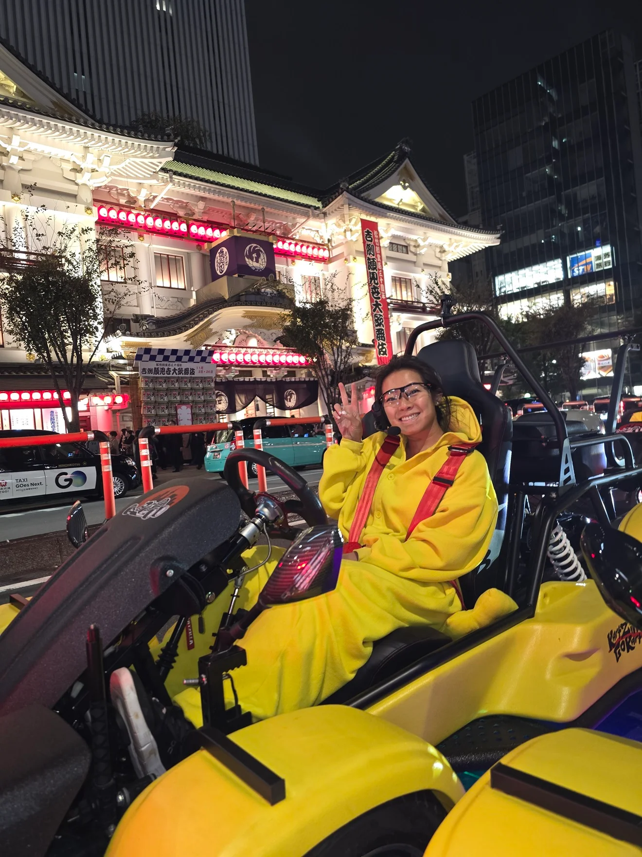 Smiling woman in a yellow costume giving a peace sign from a go-kart at night, with the ornate Kabukiza theatre illuminated behind her.