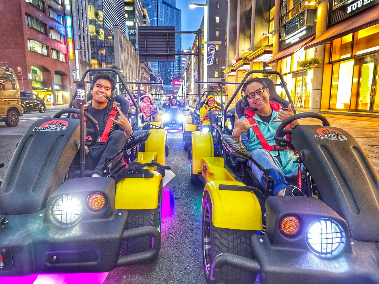 Two joyful drivers in black and yellow go-karts with vivid pink, purple, and blue underglow navigating Nihonbashi at dusk.