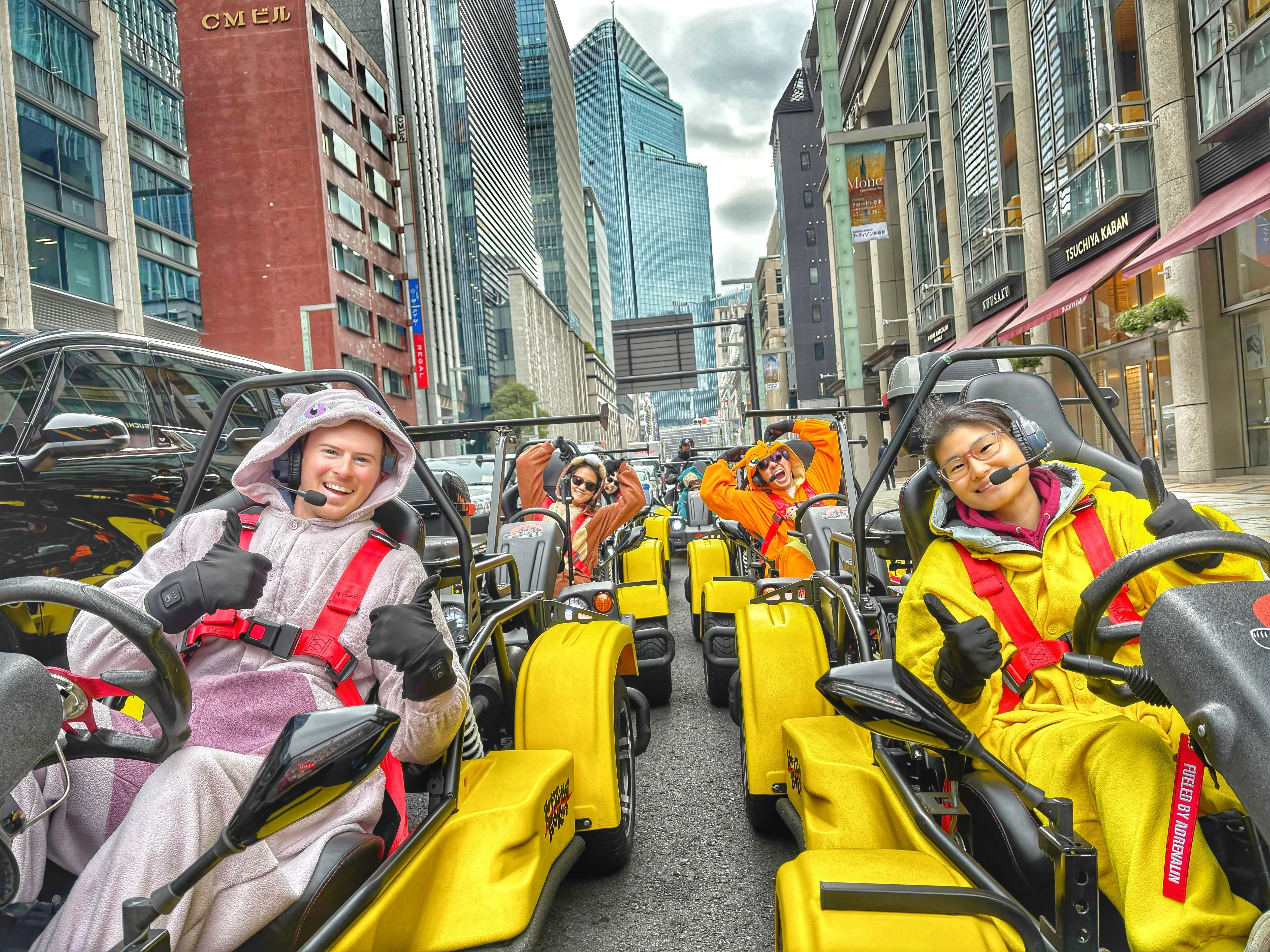 Cheerful group in vibrant yellow go-karts wearing colorful animal onesies, driving through Nihonbashi on a bright overcast day.