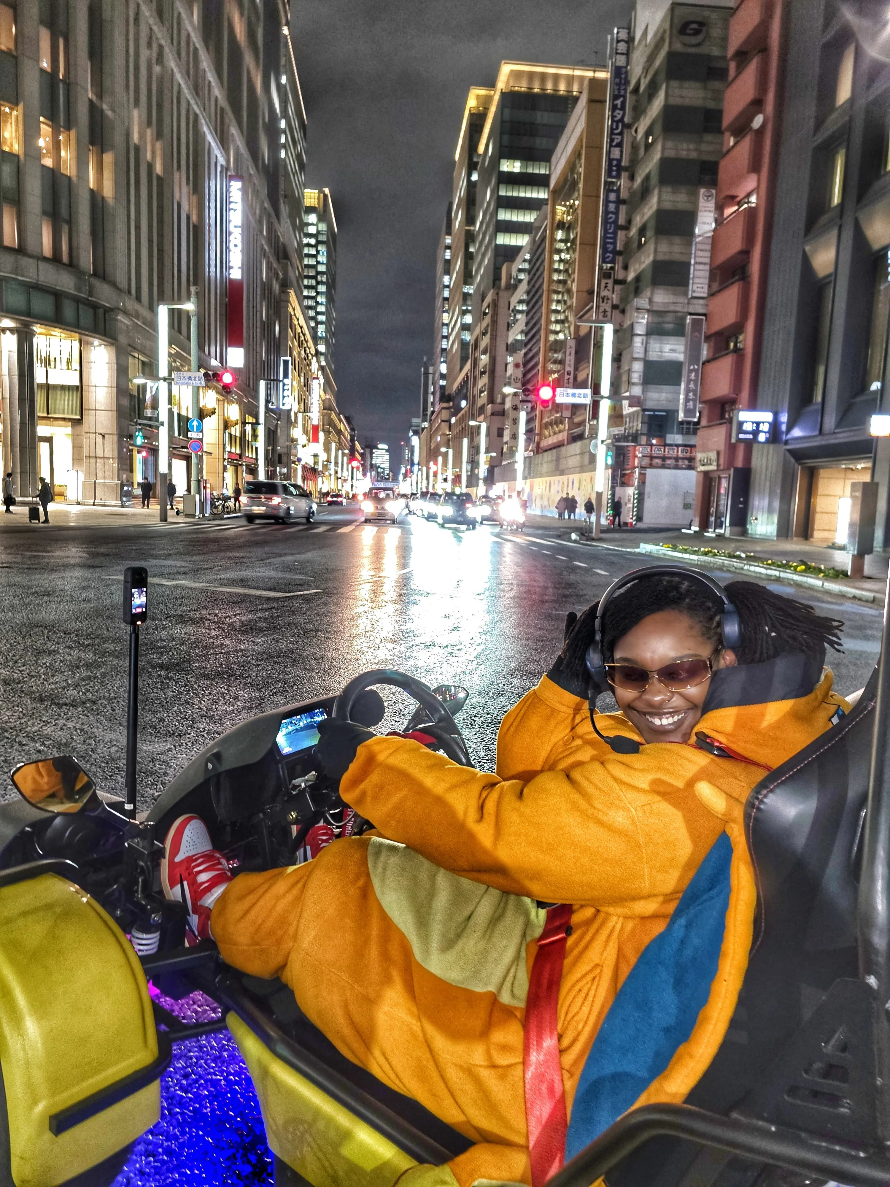 Smiling woman in a vibrant orange jumpsuit seated in a go-kart on a wet Nihonbashi street at night, with illuminated Mitsukoshi signage behind.