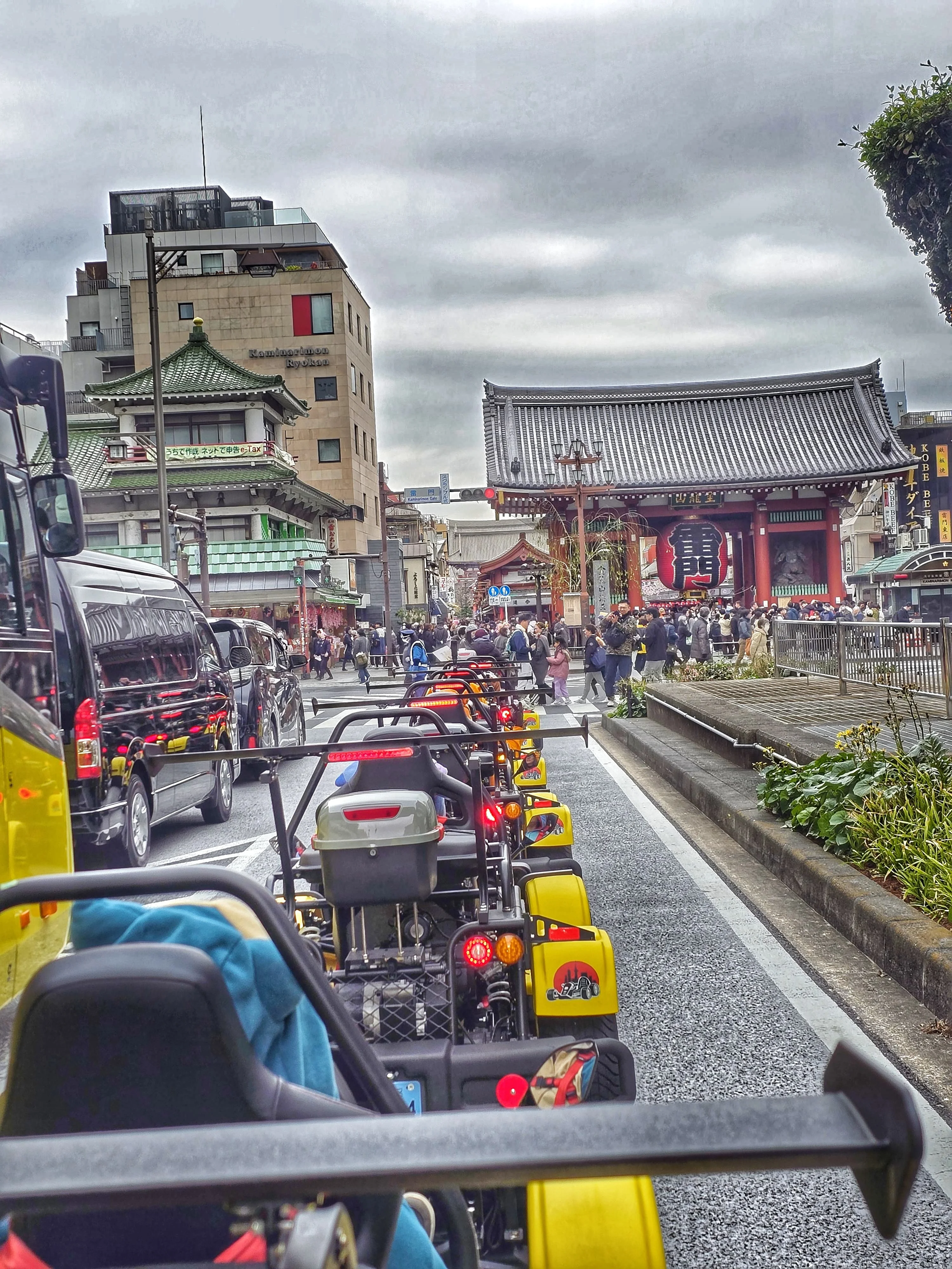 Line of yellow go-karts with glowing red taillights facing the iconic Kaminarimon Gate of Senso-ji Temple, bustling with pedestrians.