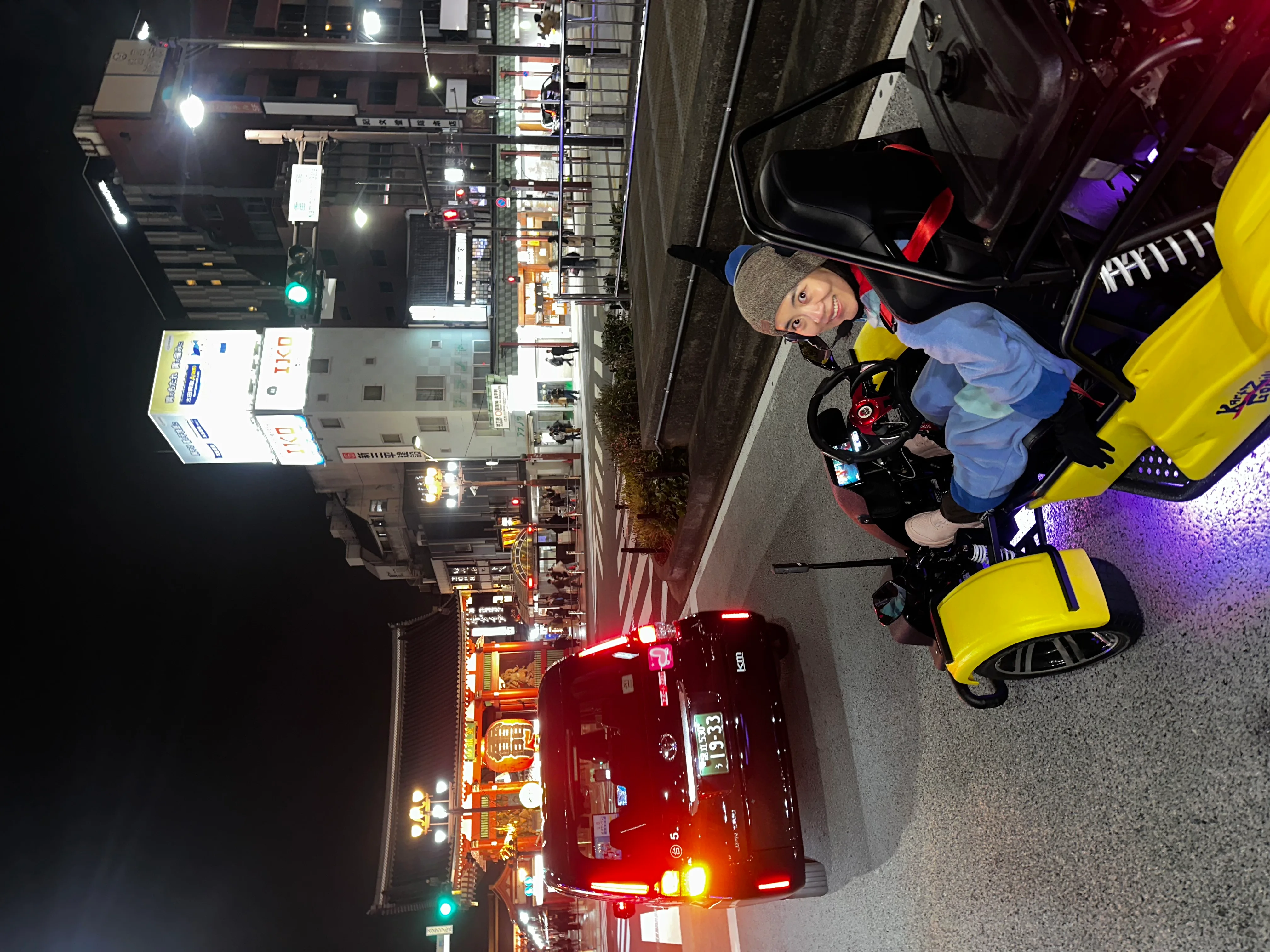 Smiling driver in a bright yellow go-kart with purple underglow, driving past the illuminated Kaminarimon Gate of Senso-ji Temple at night.