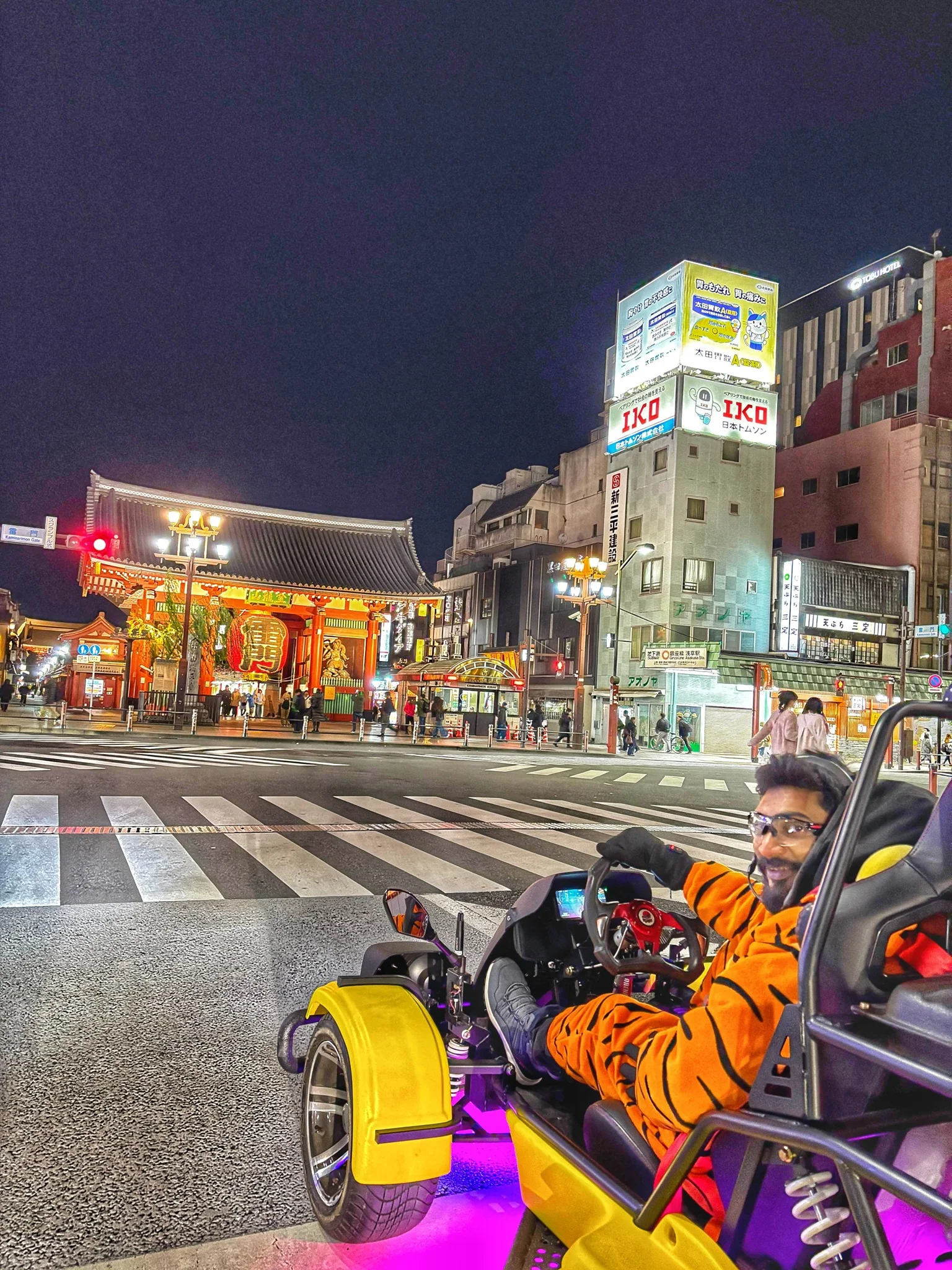 Man in a vibrant tiger-striped suit seated in a yellow kart at night, with the majestic red Kaminarimon Gate of Senso-ji Temple behind him.