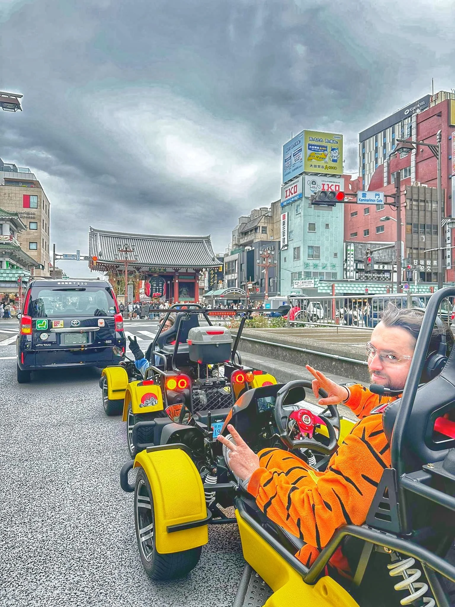 Man in orange and black tiger-print jumpsuit flashing a peace sign from a yellow go-kart, with the Kaminarimon Gate visible behind.