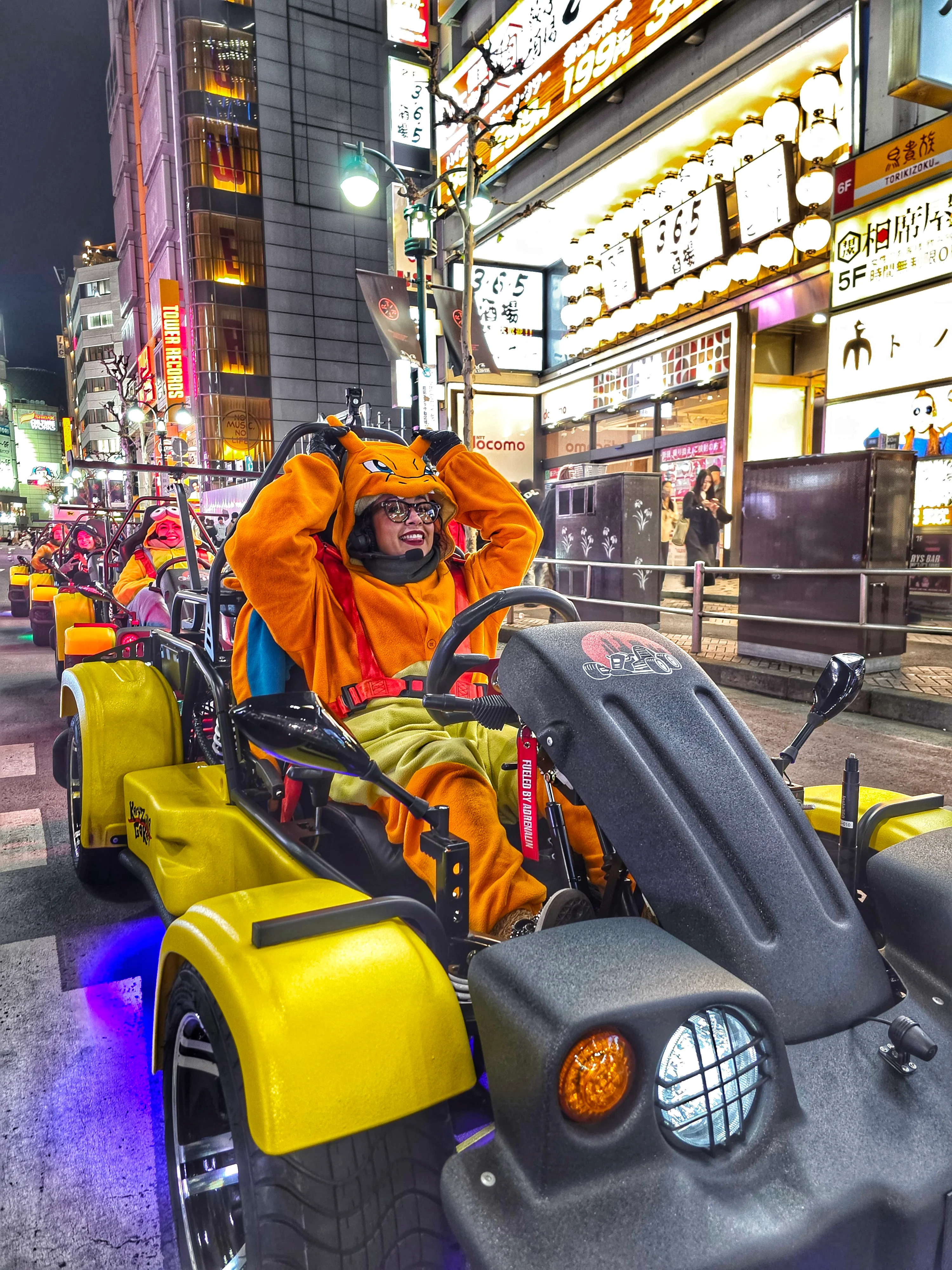Woman in a bright orange Charizard costume grinning from her yellow kart with blue underglow, navigating Shibuya at night with neon signs all around.