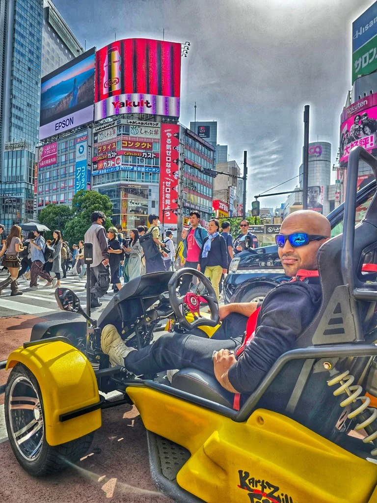 Smiling man in a vibrant yellow KartZilla go-kart on a bustling Shibuya street, with towering digital billboards and pedestrians in the background.