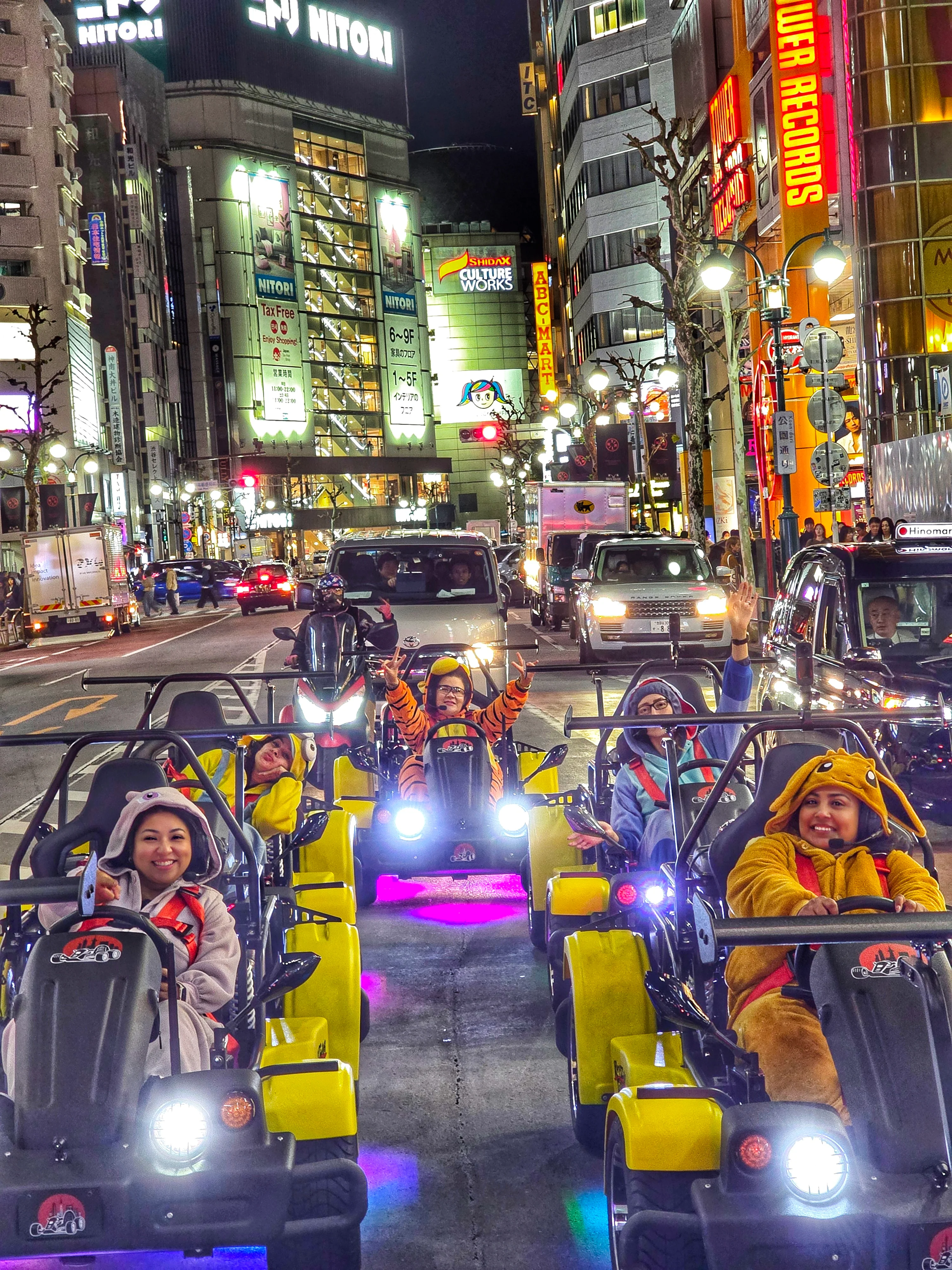 Line of yellow go-karts driven by smiling people in playful costumes through Shibuya at night, surrounded by glowing neon signs and storefronts.