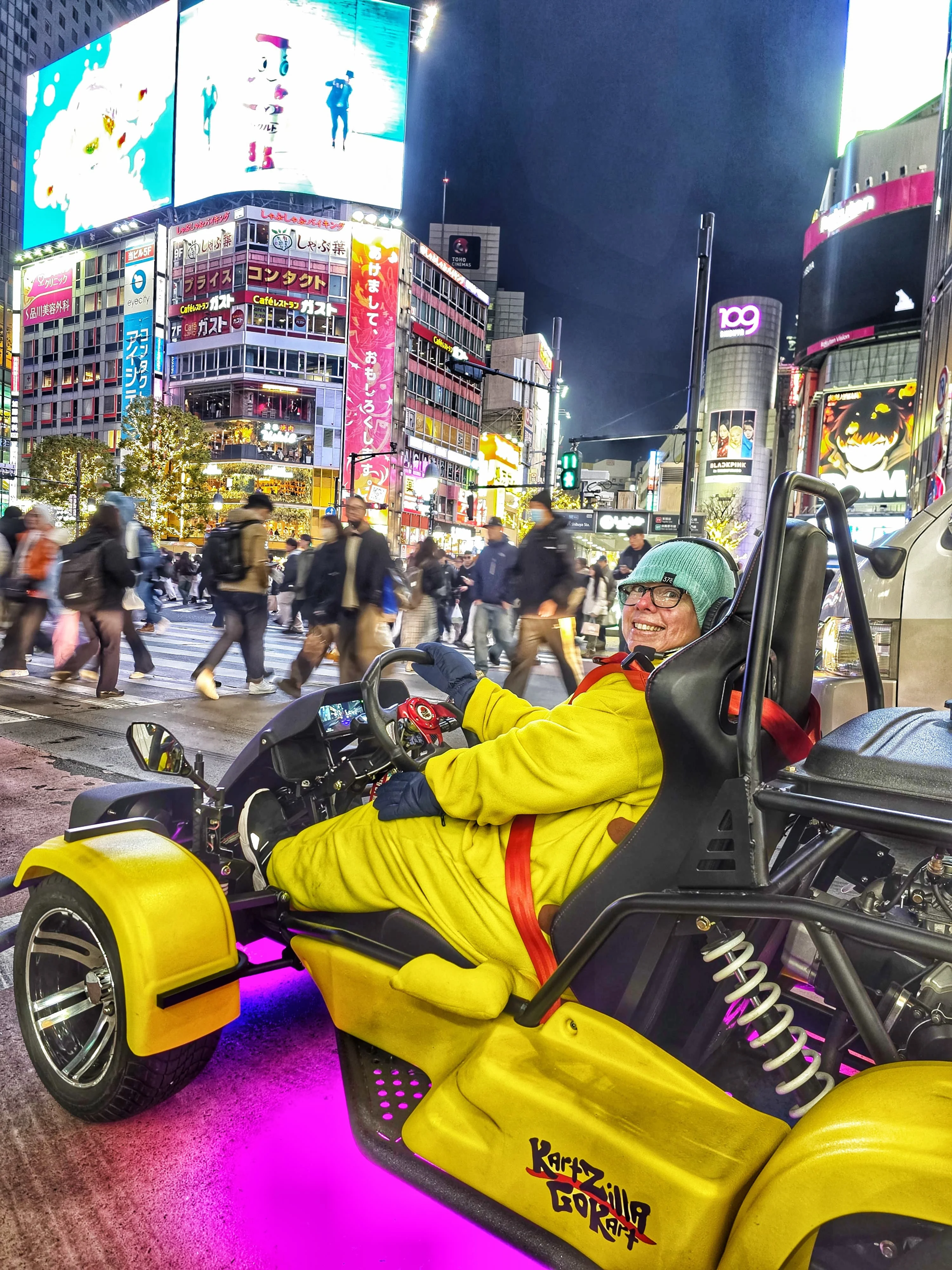 Smiling driver in a yellow jumpsuit navigating a go-kart with purple underglow through Shibuya at night, beneath the giant Rakuten Vision billboard.