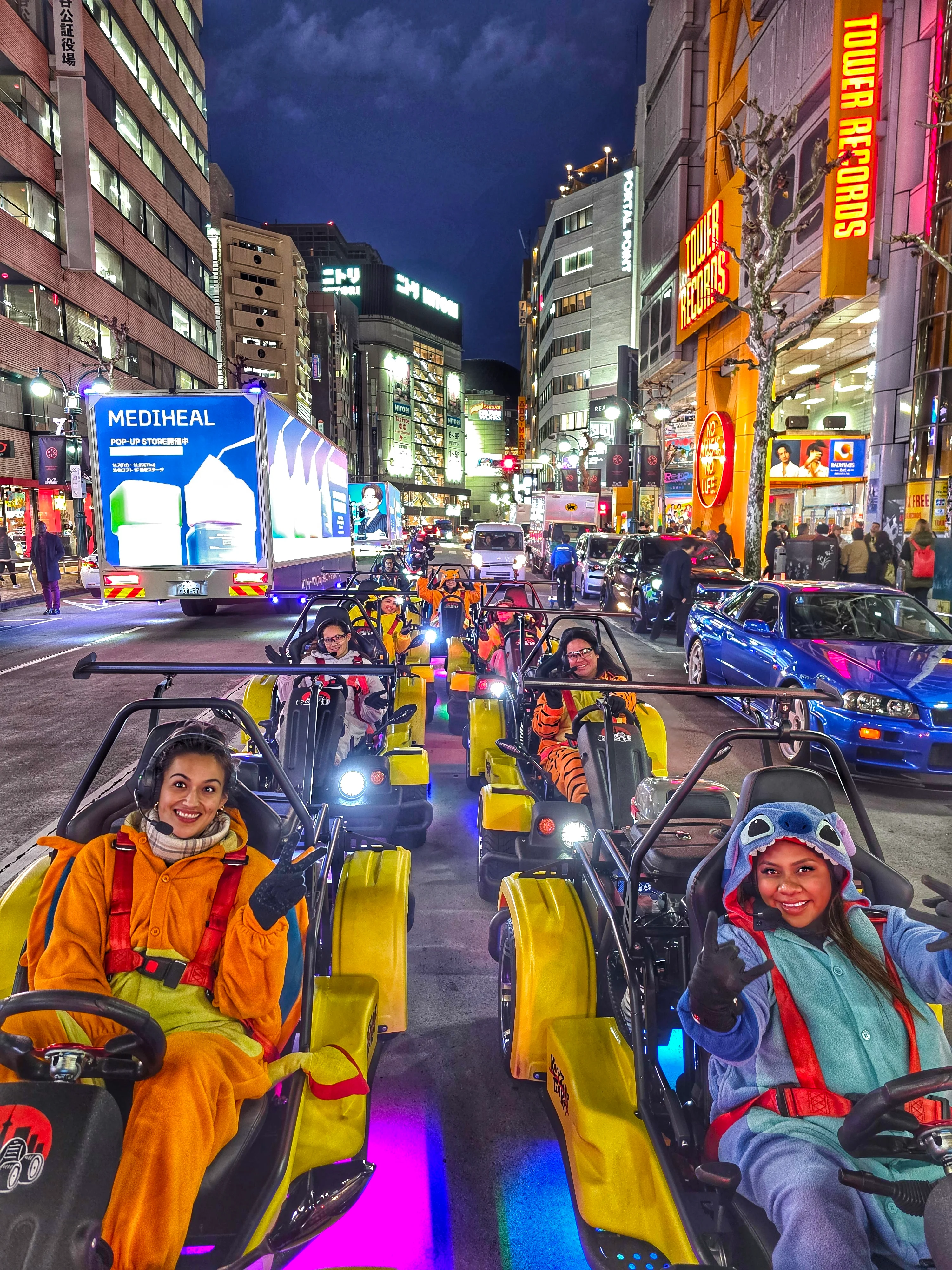 Group of smiling drivers in colorful onesies driving bright yellow karts through neon-lit Shibuya at night, with pink, blue, and purple underglow.