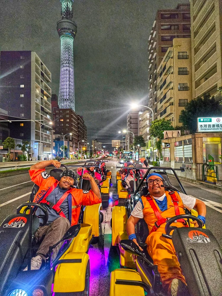 Two men in themed costumes posing in bright yellow karts at night, with the illuminated Tokyo Skytree towering in the background.