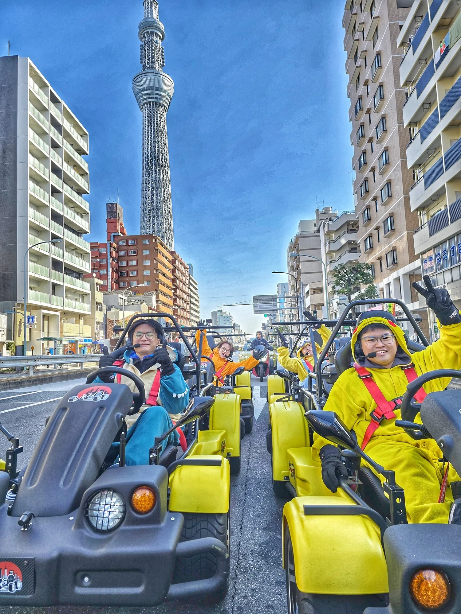 Cheerful group in a convoy of yellow and black go-karts, many in Pikachu costumes, with the iconic Tokyo Skytree towering in the background under a clear sky.