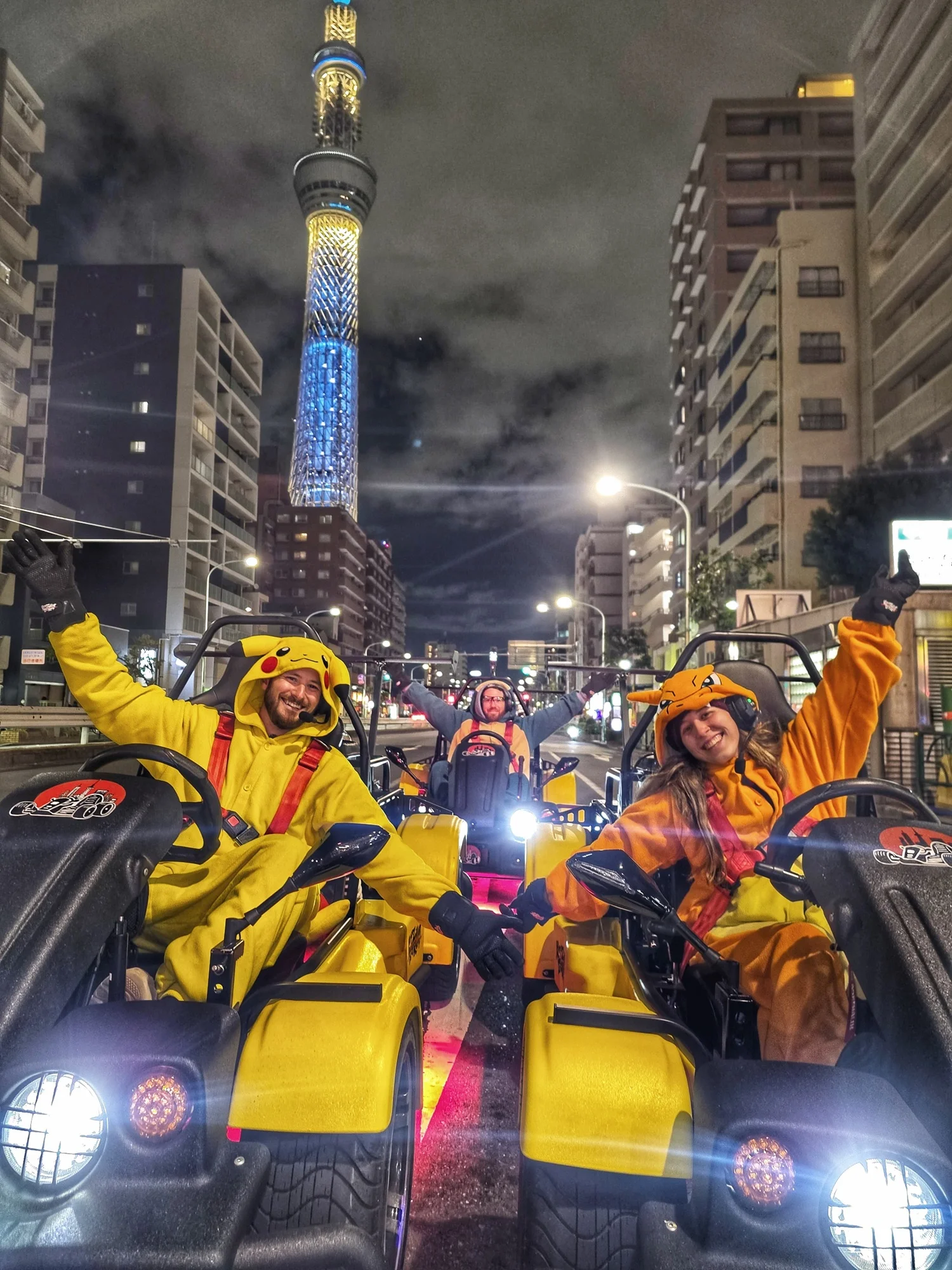 Three cheerful riders in Pikachu and Charizard costumes navigating karts through the city at night, with the glowing Tokyo Skytree visible in the distance.