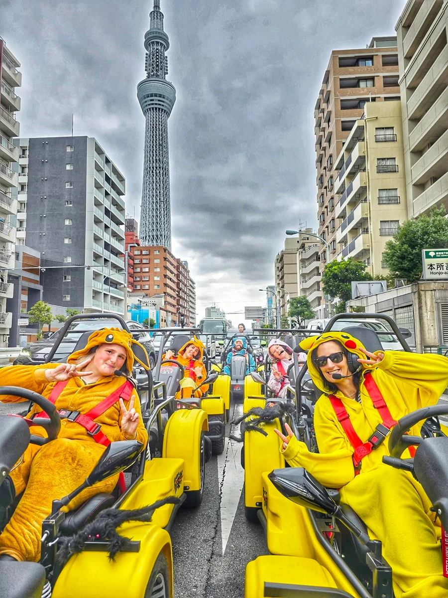 Cheerful group driving a convoy of bright yellow karts, most in Pikachu onesies, with the towering Tokyo Skytree dominating the background.