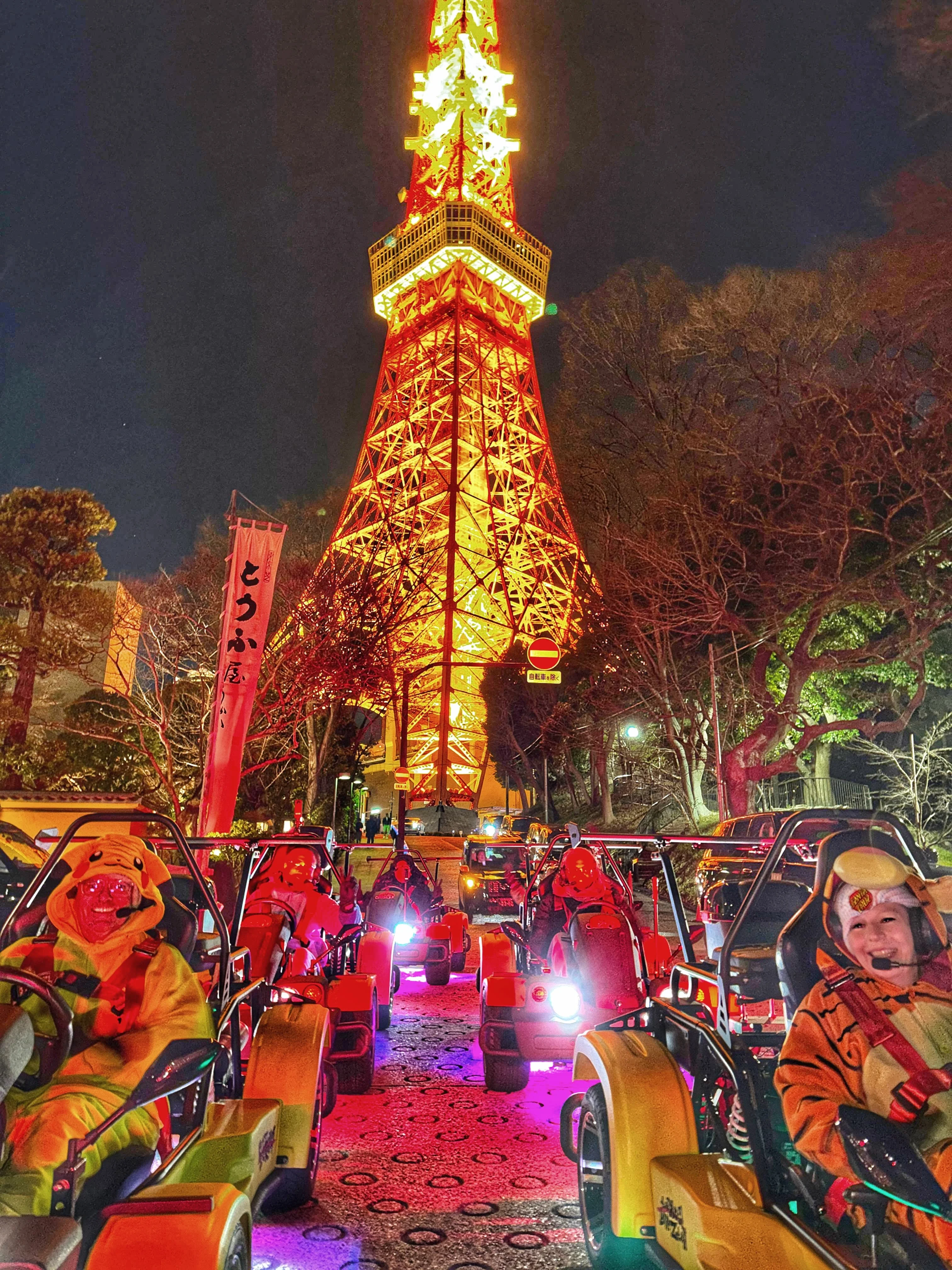 Procession of go-karts with glowing headlights and colorful underglow on a Tokyo street at night, with the illuminated Tokyo Tower behind bare trees.