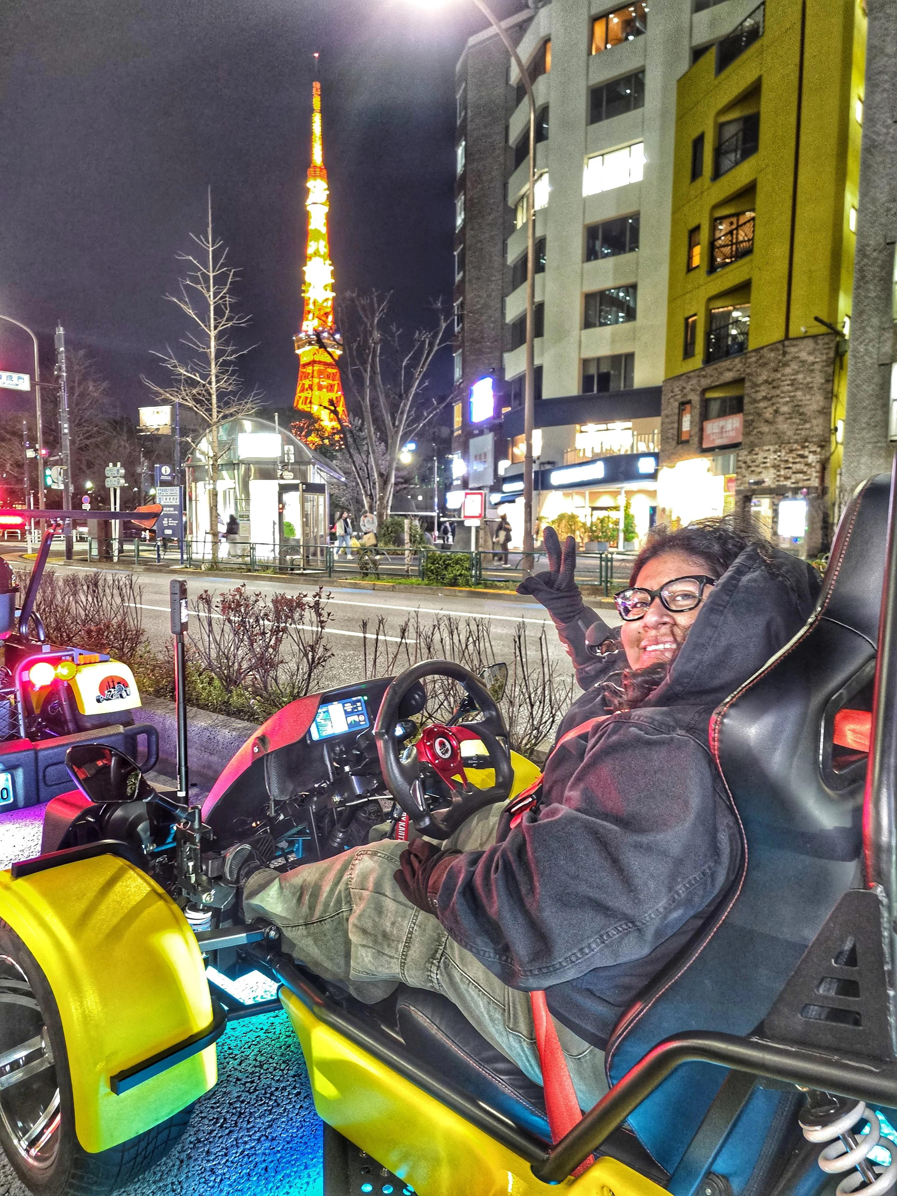 Happy driver flashing a peace sign from a bright yellow kart with blue underglow, with the glowing Tokyo Tower standing prominently in the night sky.