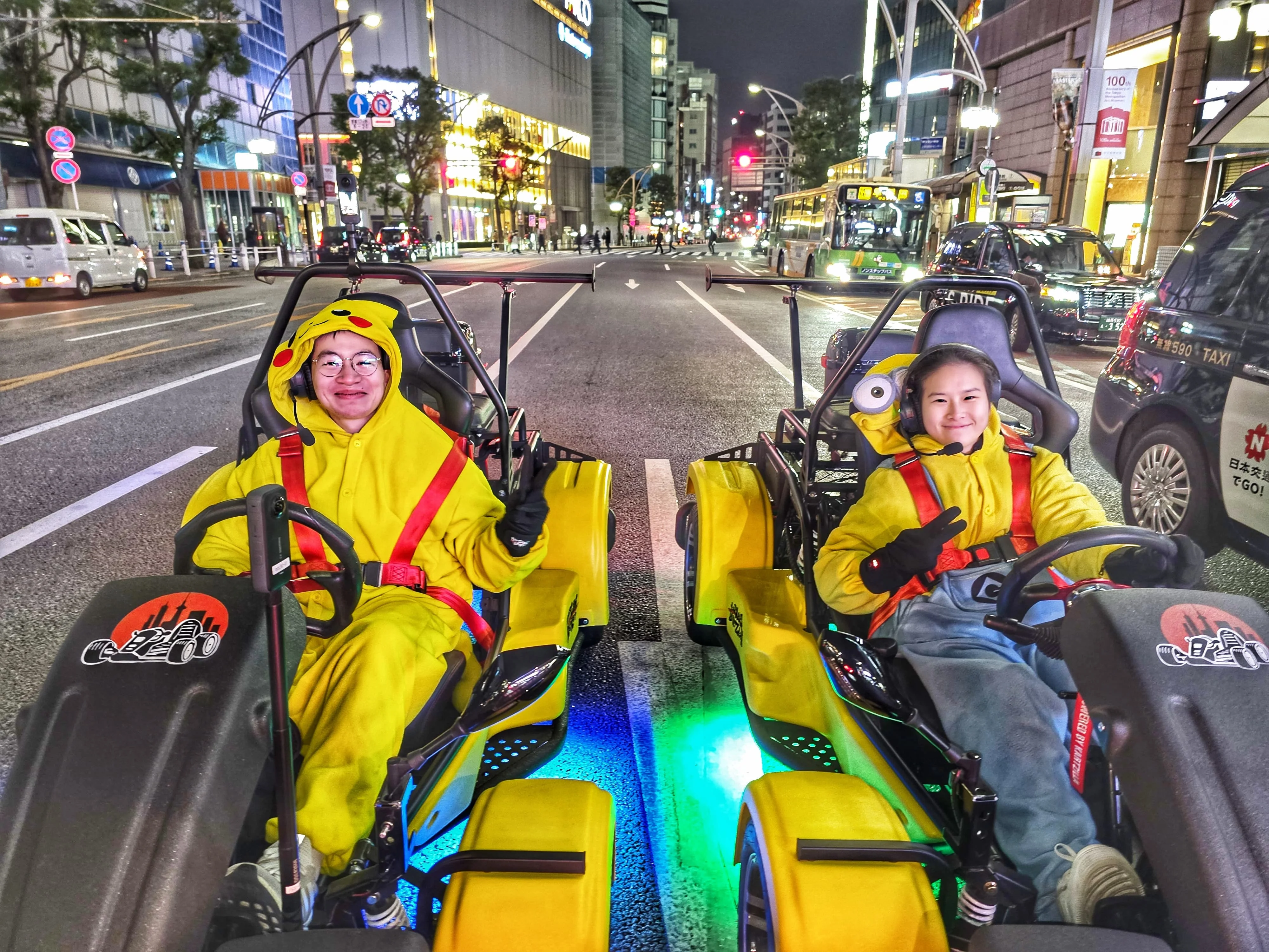 Two smiling riders in Pikachu and Minion costumes seated in yellow go-karts with blue and green underglow on a Tokyo street at night.