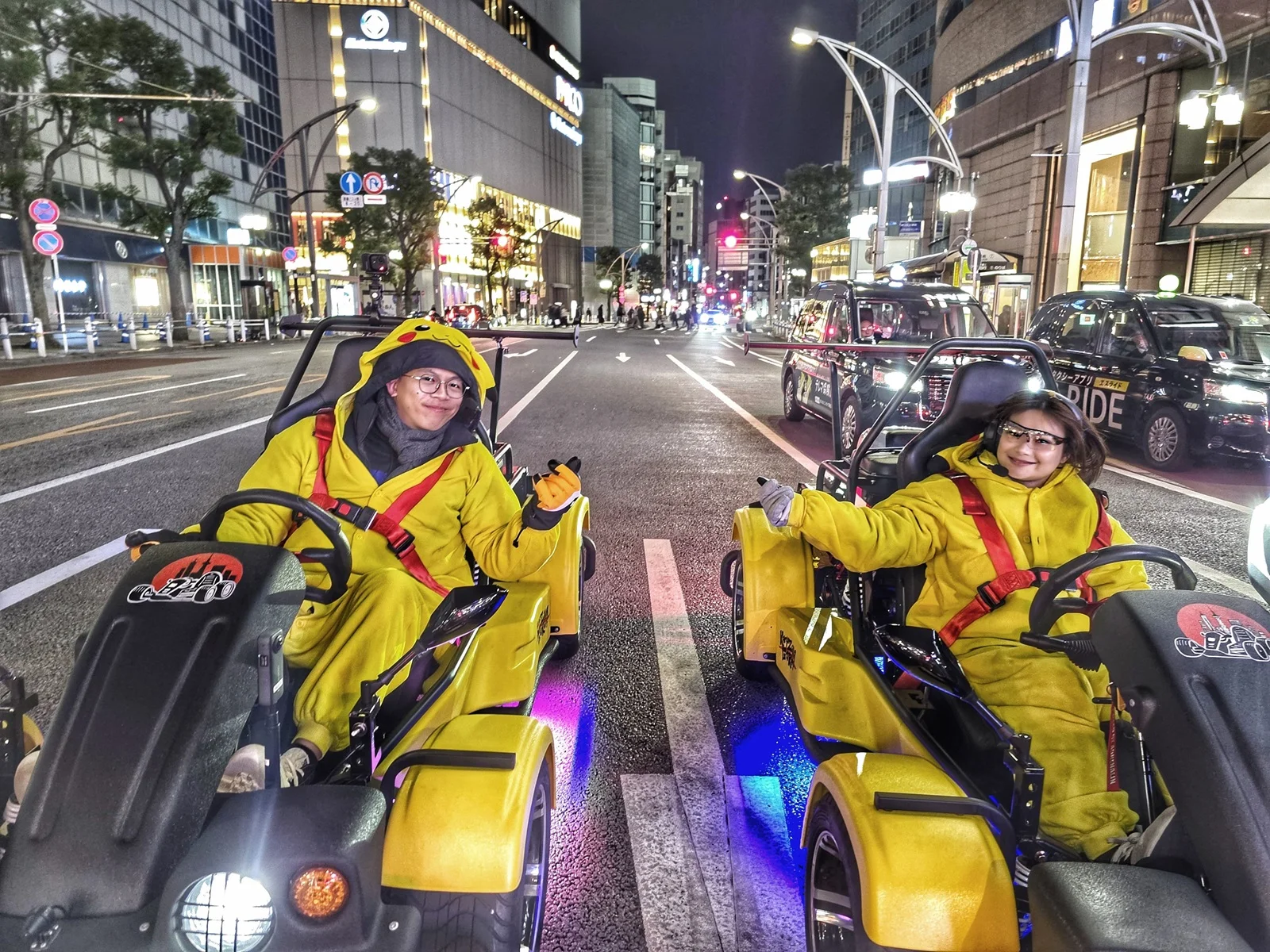 Two smiling drivers in Pikachu-themed outfits driving go-karts with bright underglow on a wet Tokyo street at night, with city lights reflecting off the asphalt.